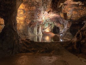 Photographie des Mines d'Argent des Rois Francs, montrant un vaste intérieur de grotte avec des lumières artificielles mettant en évidence les textures et les nuances de la roche. L'eau calme du lac souterrain reflète la lumière, ajoutant de la profondeur et de la sérénité au paysage souterrain historique